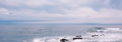Ocean waves breaking against a rocky shoreline beneath an overcast sky.